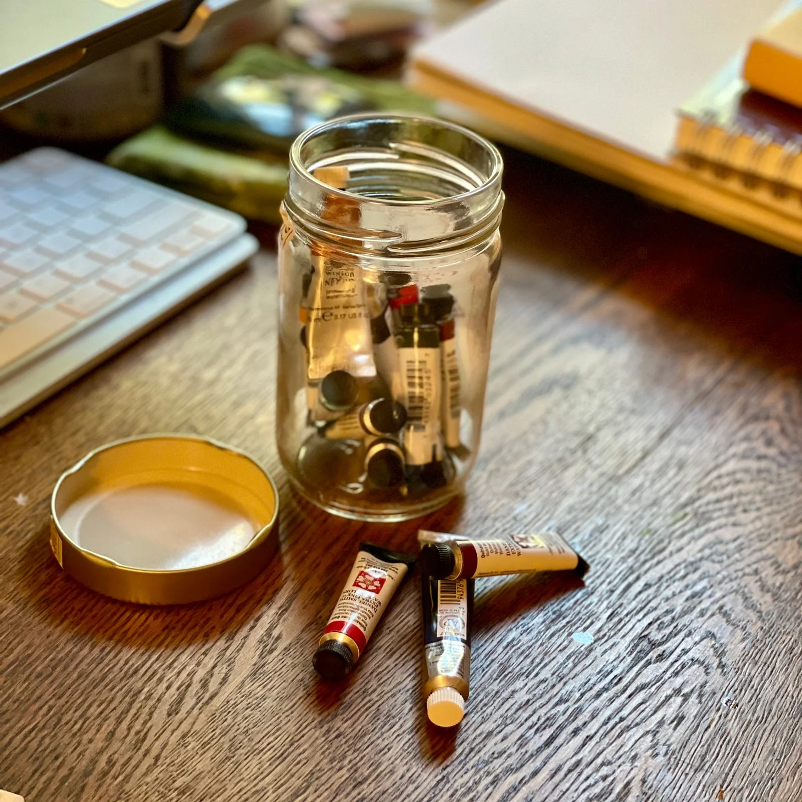 Tubes of watercolor paint in a glass jar, on a desk.