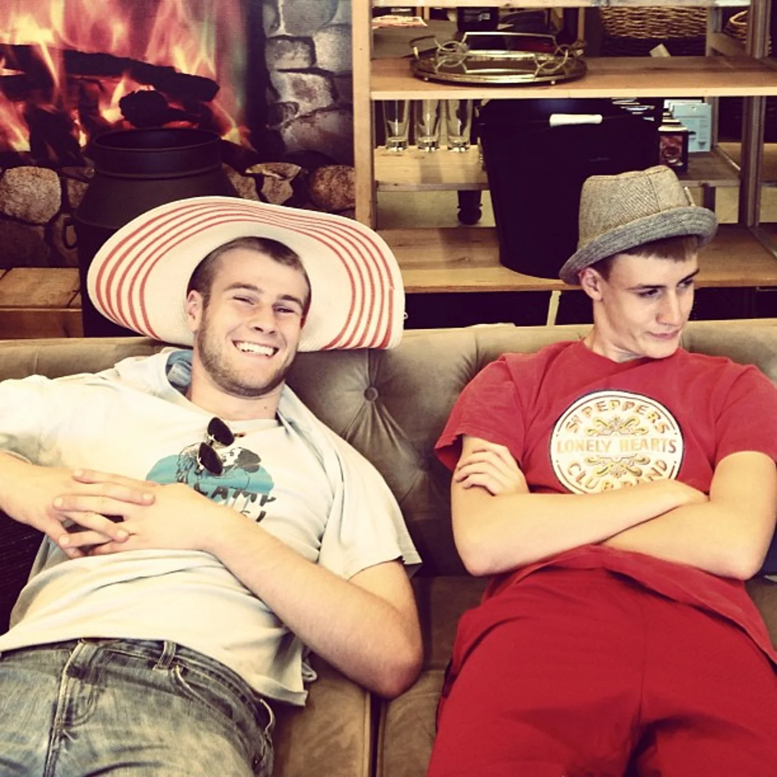 Two teenagers wearing silly hats sit on a couch at a furniture store. The boy on the right is grinning at the camera. The boy on the left is looking away in mock disdain.